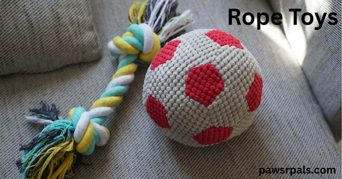 Rope Toy. Green, yellow and white material entwined and knotted at each end next to a white and red small football shaped knitted ball sat on a grey surface